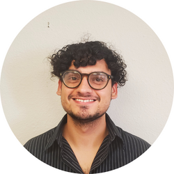 Close-up headshot of Samuel, with curly hair and glasses, standing in front of a white wall.